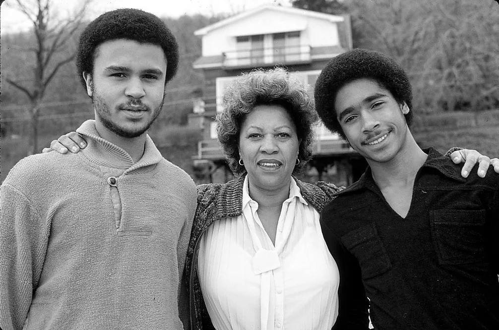 Photograph of Toni Morrison standing between her two grown sons, who have their arms around her, outside a house among bare trees.