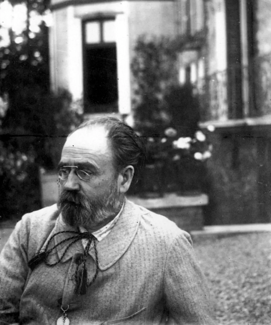 Photograph of Émile Zola in later life, bearded, in pince-nez and a light jacket, sitting in front of his house at Médan.